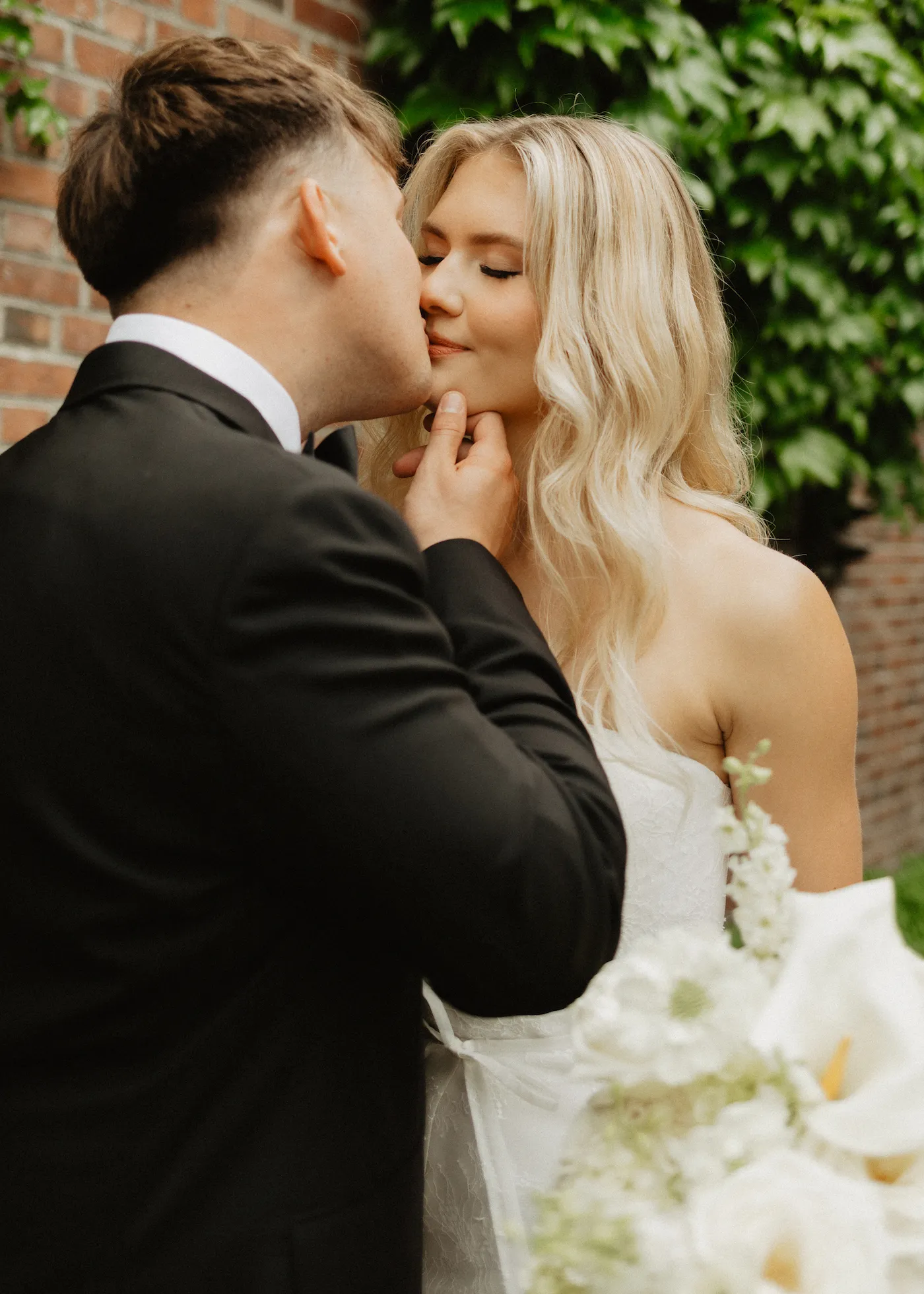 Wedding portrait of a bride and groom kissing beside an ivy-covered brick wall